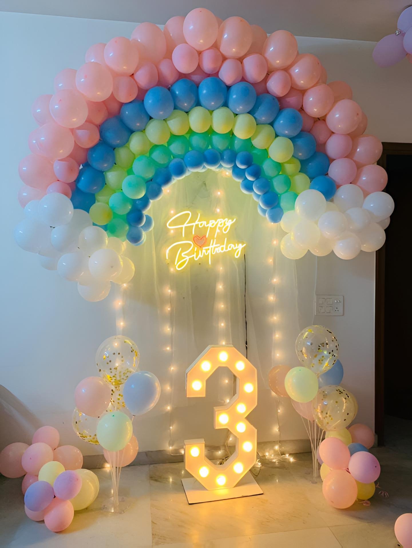Pastel rainbow birthday decoration with colourful balloon arch, white balloon clouds, Happy Birthday LED neon light, illuminated age number, warm fairy lights and pastel balloon bunches for a kids birthday celebration in Delhi NCR.