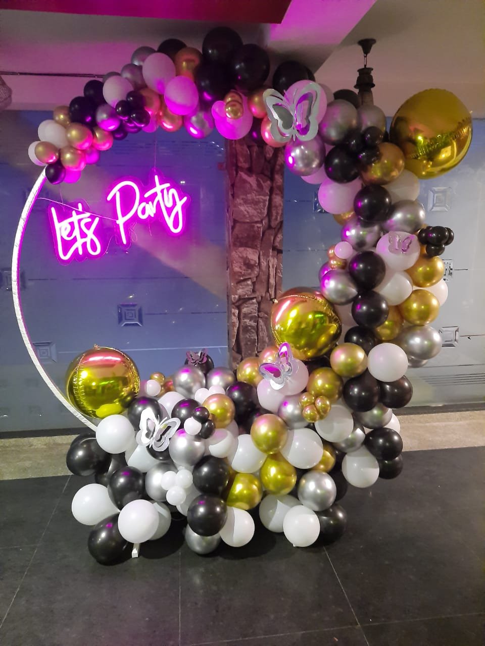 Let's Party ring balloon decoration featuring an elegant ring backdrop, organic balloon garland in black, white, gold chrome and silver, 4D foil balloons, large chrome balloons, 3D decorative butterflies and a glowing Let's Party LED neon light. Stylish party decoration setup for birthdays, anniversaries and special celebrations in Delhi NCR